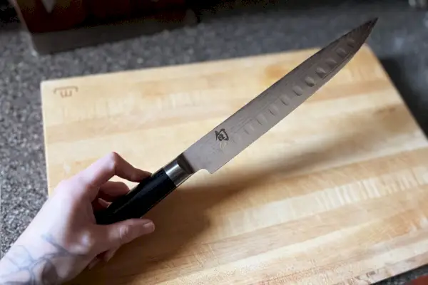 Una persona que sostiene el cuchillo Shun DM-0720 Classic de 9 pulgadas con punta hueca sobre una tabla de cortar